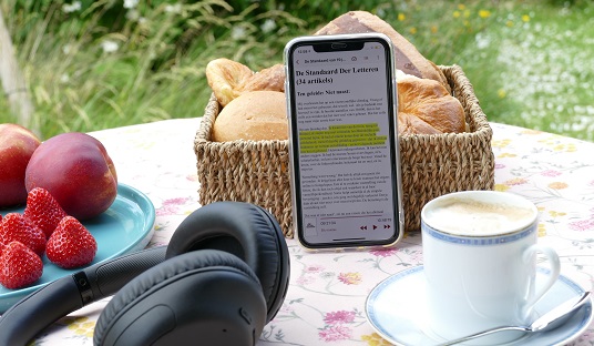 Een smartphone staat op de ontbijttafel. Op de smartphone is de AndersLezen-app geopend; een gele balk markeert de tekst die voorgelezen wordt.
