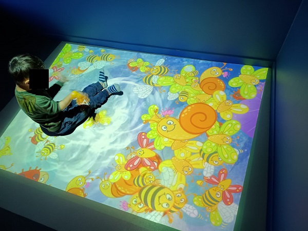 Een kind zit op de vloer, waarop een vloerprojectie met getekende bijen, slakken en vlinders te zien is.