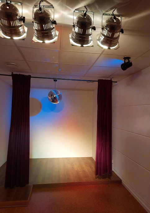 Een klein podium wordt verlicht door vier schijnwerpers. Boven het podium hangt een discobal, beschenen door blauw licht, wat blauwe lichtpuntjes reflecteert op de muren.